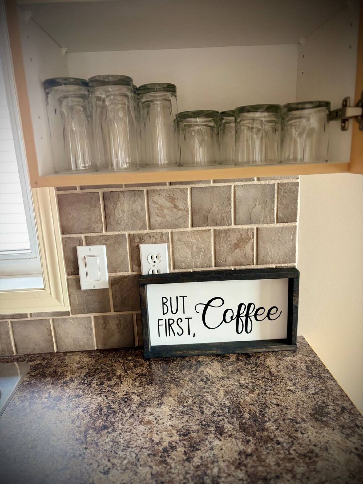 A kitchen counter with a framed sign that reads "BUT Coffee FIRST," above jars on a shelf.