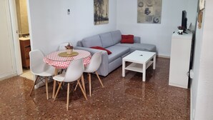 Un espace de vie compact comprenant un canapé gris, une petite table ronde avec des chaises, une table basse blanche et un ventilateur de plafond.