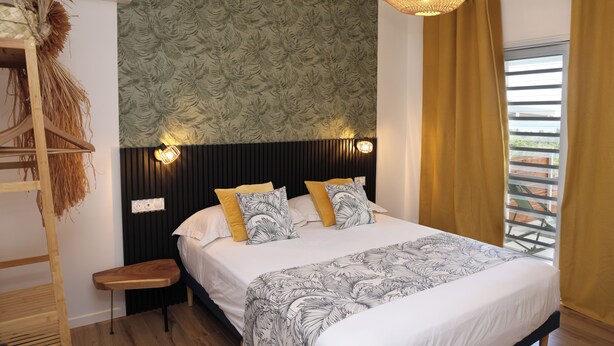 Slaapkamer met een bed, nachtkastje, wandlamp, raam met gordijnen en een muur met een bladerpatroon.