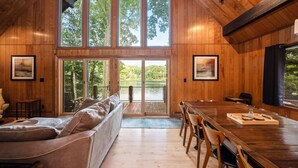 Un intérieur en bois avec une grande table à manger, un canapé et une vue sur une terrasse extérieure, accessible par des portes vitrées.