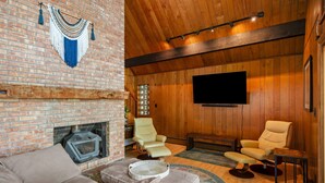 Un salon chaleureux avec une cheminée en briques, deux fauteuils en cuir, un téléviseur à écran plat et un plafond en bois.