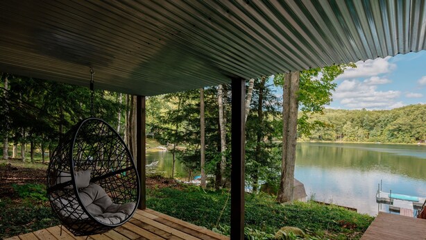 Une terrasse en bois avec un fauteuil suspendu donnant sur un lac, le tout entouré d’arbres.
