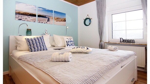 A neatly made bed with a striped bedspread, pillows, and a rolled towel. There are two wall-mounted lamps, a small decorative item, and a window with curtains. - Reihenhaus Land and Sea in Busum (Westerdeichstrich)