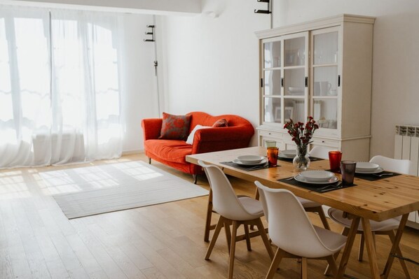 Un coin repas comprenant une table en bois, des chaises blanches, un canapé rouge et un meuble blanc avec des portes en verre.