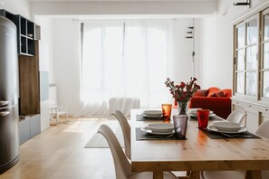 Ein Essbereich mit Holztisch, weißen Stühlen, einem roten Sofa und einem großen Fenster mit durchsichtigen Vorhängen.
