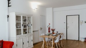 Een eetruimte met een houten tafel en witte stoelen, een wit kastje met glazen deuren, en een rode brandblusser aan de muur.