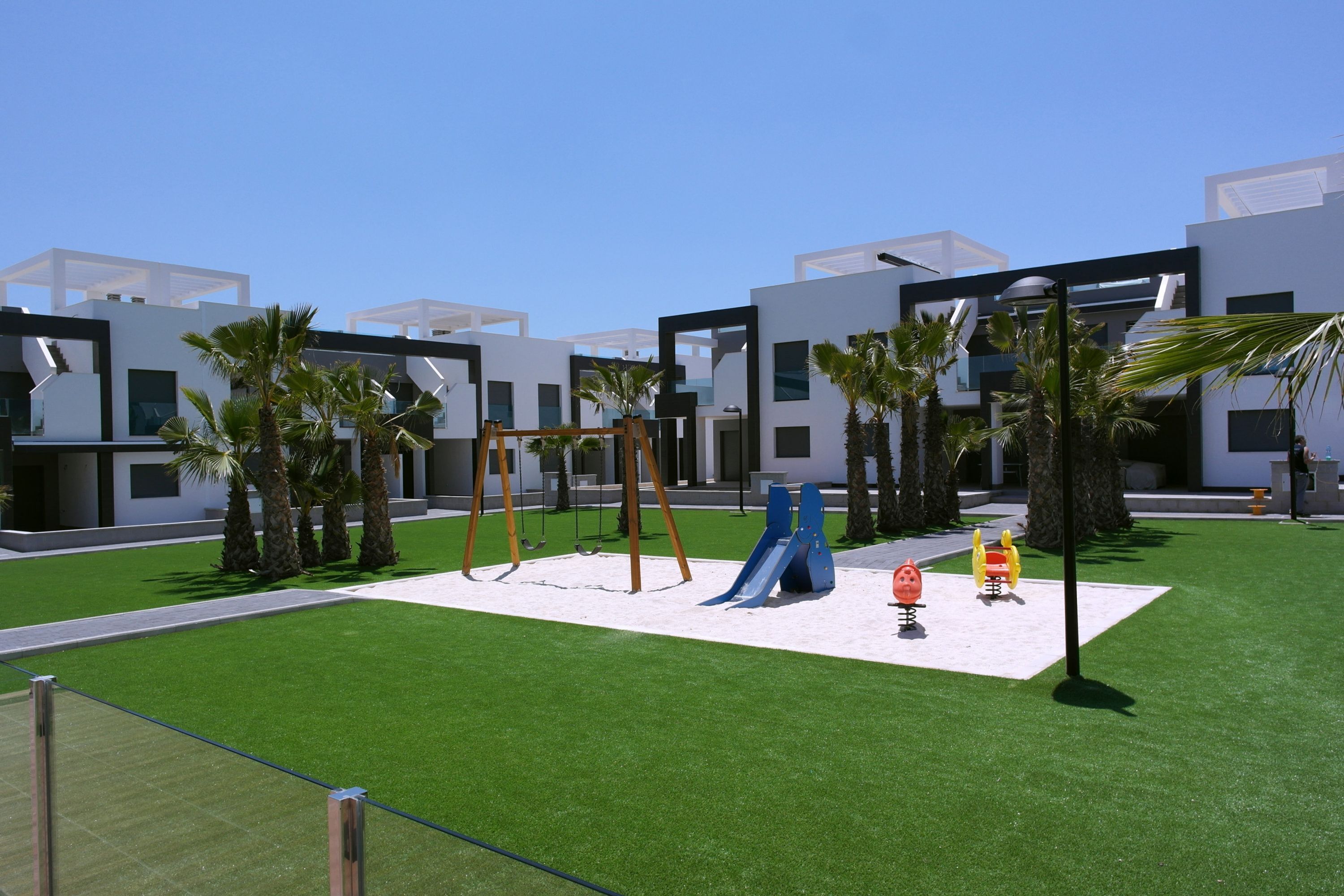 Un parque infantil con columpios y toboganes, rodeado de palmeras y edificios modernos.