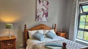Een slaapkamer met een houten bed, een nachtkastje, een lamp, een raam met gordijnen en een bloemistisch schilderij aan de muur.