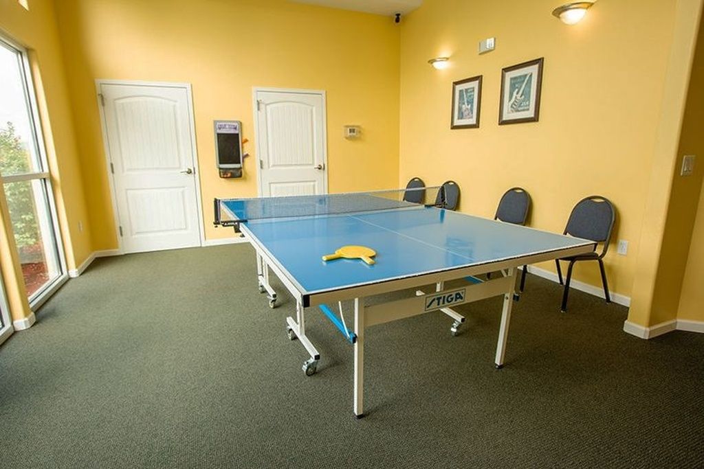 Una mesa de ping pong en una habitación con paredes amarillas, dos puertas blancas y un ventanal.