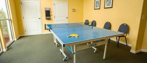 Una mesa de ping pong en una habitación con paredes amarillas, dos puertas blancas y un ventanal.