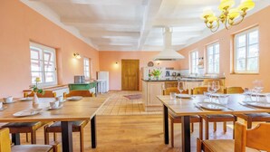 A spacious dining area with wooden tables and chairs, a kitchen in the background, and large windows allowing natural light. - Vacation in the castle up to 15 pers. Apartment: Burggraf & Himmelsleiter (Großbodungen)