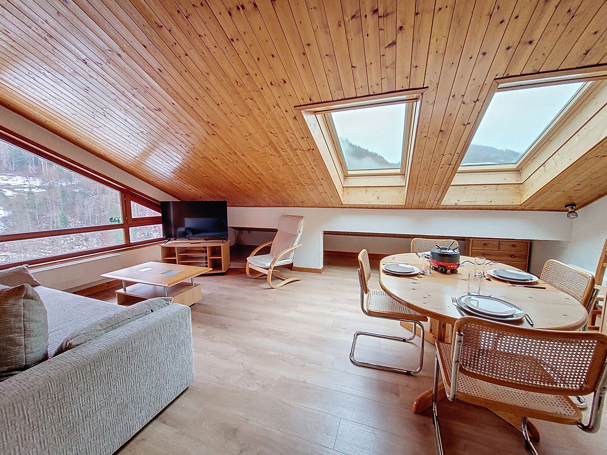 Um amplo loft com pé direito alto, vigas de madeira, uma televisão grande, área de refeições e vista para o exterior.