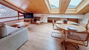 A spacious loft with high ceilings, wooden beams, a large TV, a dining area, and a view of the outdoors.