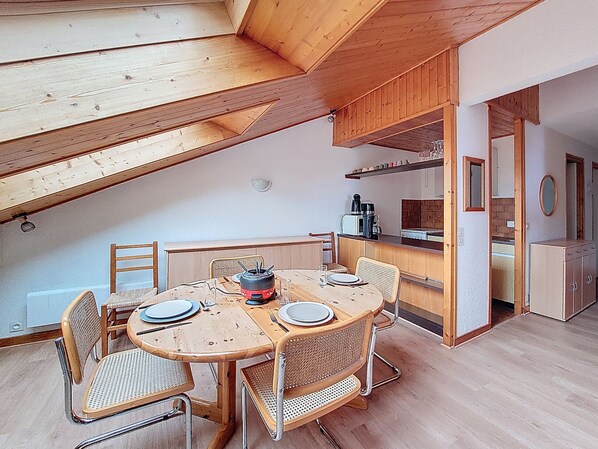 A dining area with a wooden table and chairs, a kitchen in the background, and a loft space above.