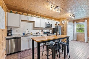 Una cocina con armarios blancos, encimera de madera, lavavajillas de acero inoxidable, microondas, refrigerador y una zona de bar con taburetes.