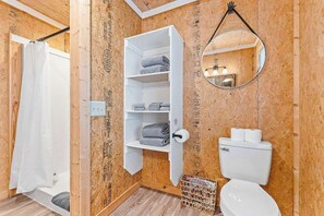 A bathroom with a wooden wall, a white shelf with towels, a toilet, and a mirror. - Hot Tub, Fire Pit, Pet Friendly Close to Lake (Eureka Springs)