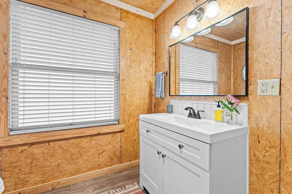 A bathroom with wooden walls, a white vanity, a mirror with lights, and a window with blinds.
