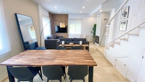 A modern living room with a wooden dining table, dark chairs, a sofa, a TV, and a staircase.