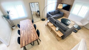 A modern living room with a wooden dining table, grey sofa, and a TV mounted on the wall.