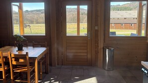 Un interior de madera con una mesa de comedor, sillas y una planta en maceta. Se ven dos ventanas que ofrecen vistas a un paisaje rural.