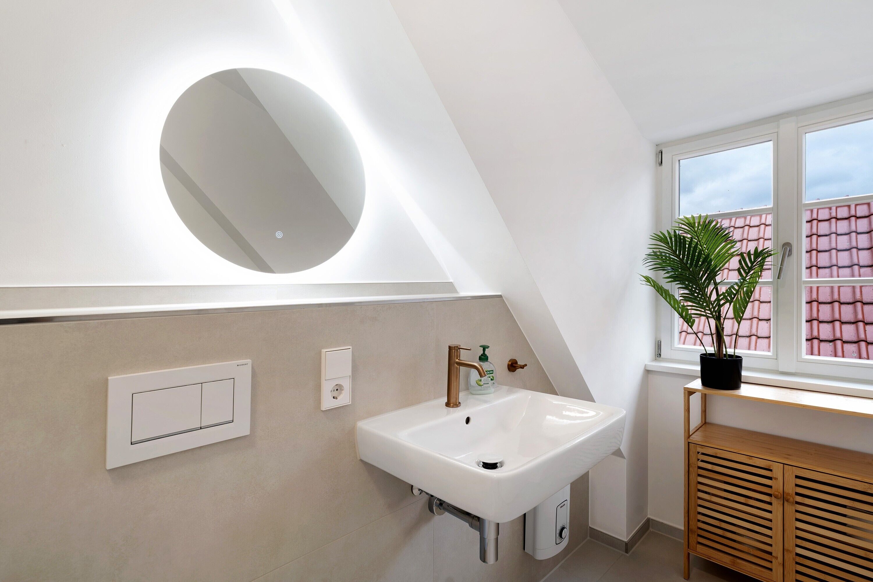 Ein modernes Badezimmer mit einem weißen Waschbecken, einem runden Spiegel, einer Topfpflanze und einem Fenster mit Blick auf ein rosafarbenes Gebäude.