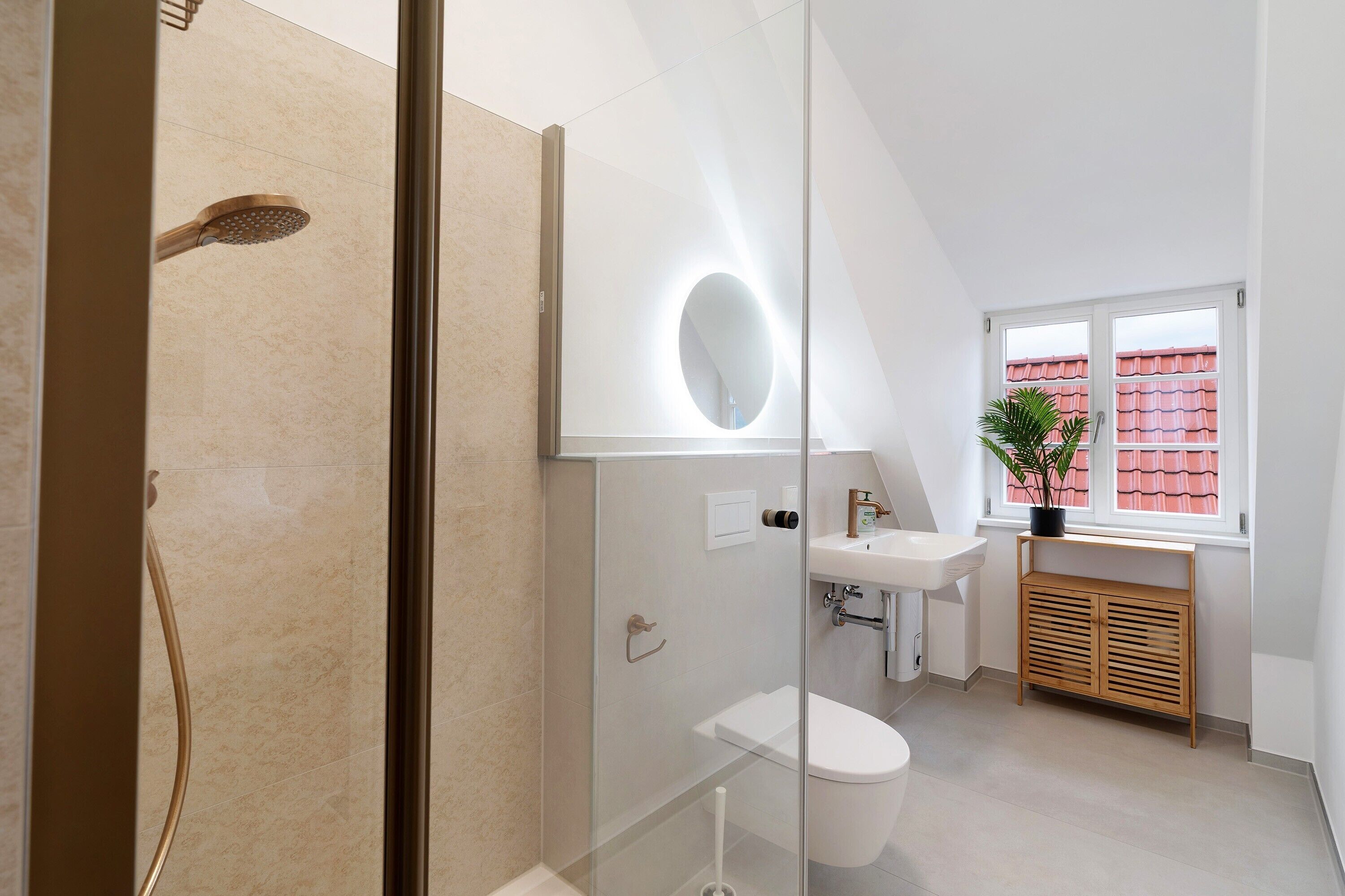 Un baño moderno con cabina de ducha de vidrio, inodoro, lavabo y ventana con vistas a edificios de tejados rojos.