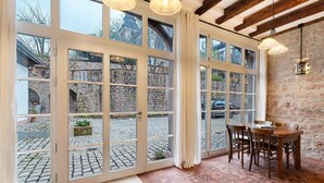 A room with large windows, a stone wall, a wooden table with chairs, and a patterned floor.