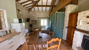 A rustic kitchen with a stone wall, wooden beams, a dining table with chairs, a microwave, a refrigerator, and a fireplace.