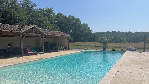 A rectangular swimming pool with clear water, surrounded by a tiled deck. There are lounge chairs and a covered seating area with a pergola roof.