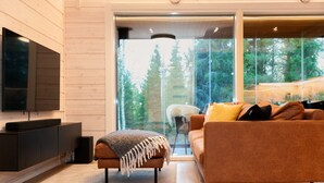 A modern living room with a large window, a flat-screen TV, a brown sofa, and a small ottoman. - Modern 3BR Villa Retreat in Ruka with Private Sauna & Near Ski Slopes (Ruka)