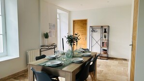 A dining area with a wooden table set for four, surrounded by chairs. There is a bookshelf with books and decorative items, and a potted plant on a small table. - Le gite de Fontiville near the Loire castles (6 to 8 people) (Veigné)