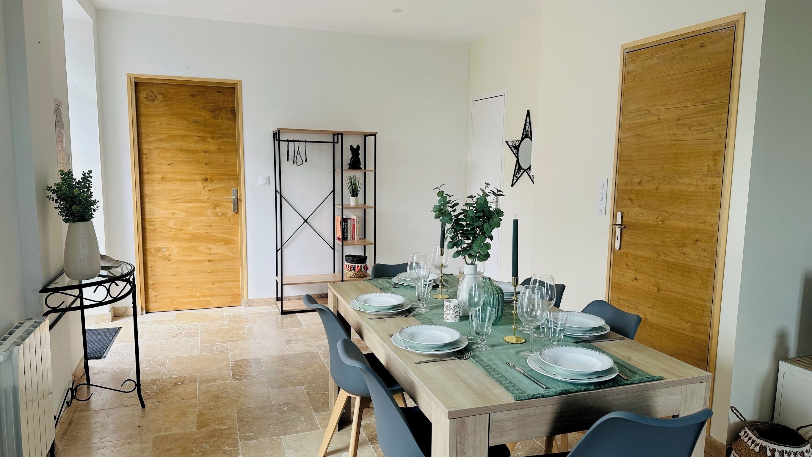A dining area with a wooden table set for four, flanked by two wooden doors, a black metal shelf unit, and a potted plant.