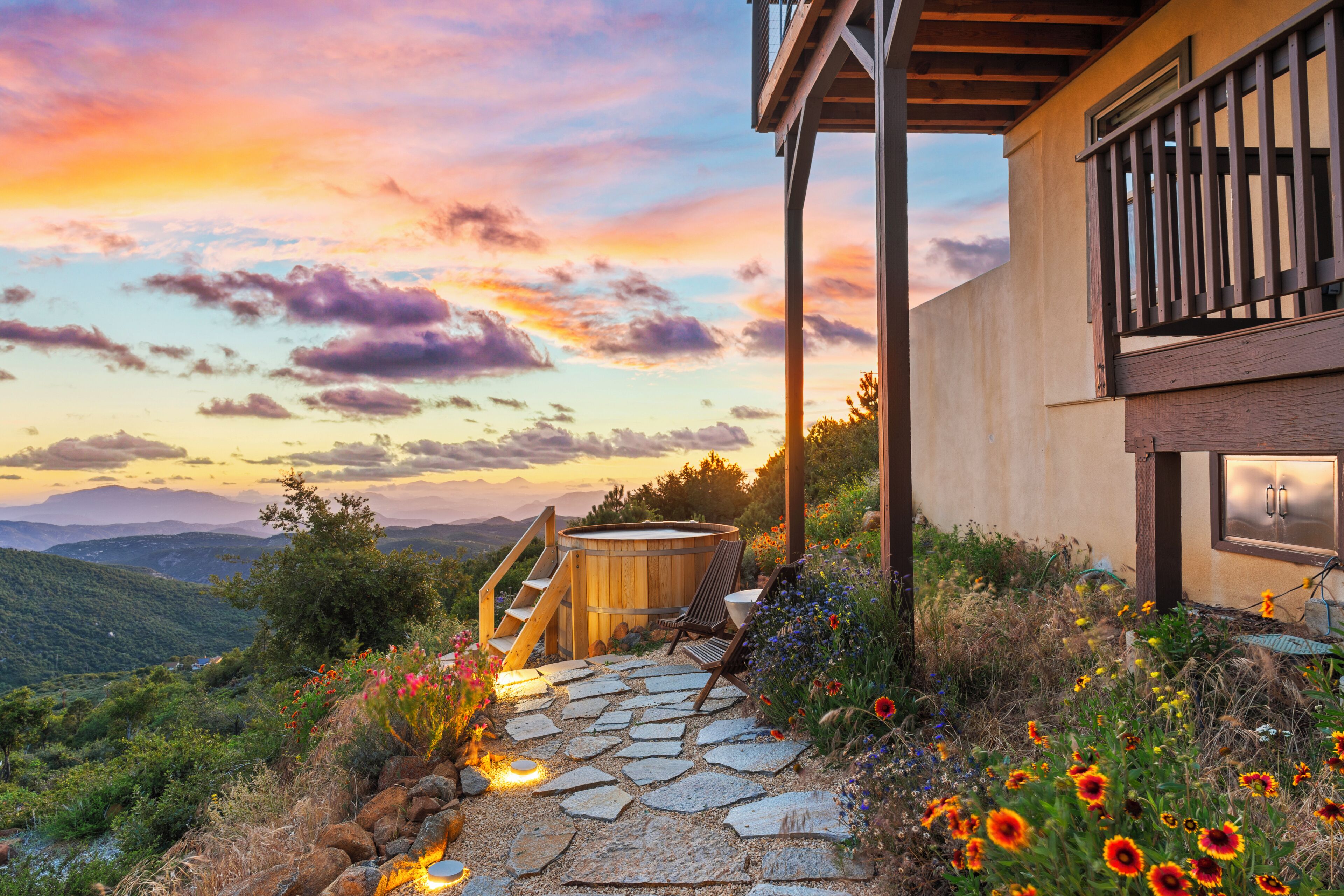 A stone pathway leading to a wooden hot tub with a view of a mountainous landscape and a sunset.