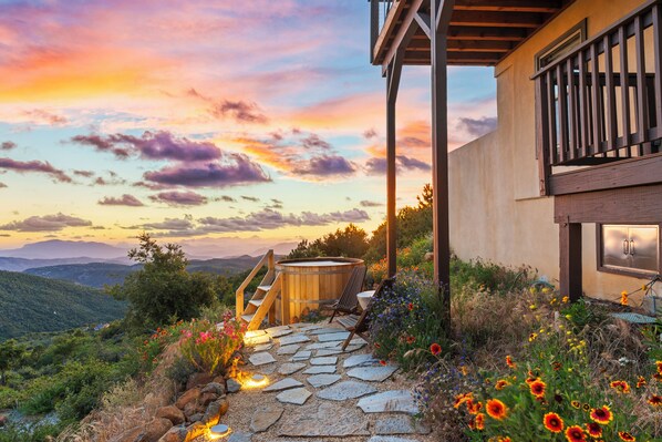 A stone pathway leading to a wooden hot tub with a view of a mountainous landscape and a sunset.