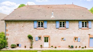 Une maison à deux étages, avec un toit recouvert de tuiles, de nombreuses fenêtres dotées de volets et un petit jardin à l’avant.