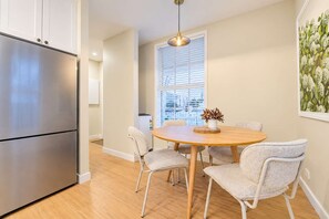 A modern kitchen with a stainless steel refrigerator, a round wooden dining table with four chairs, and a window with blinds.