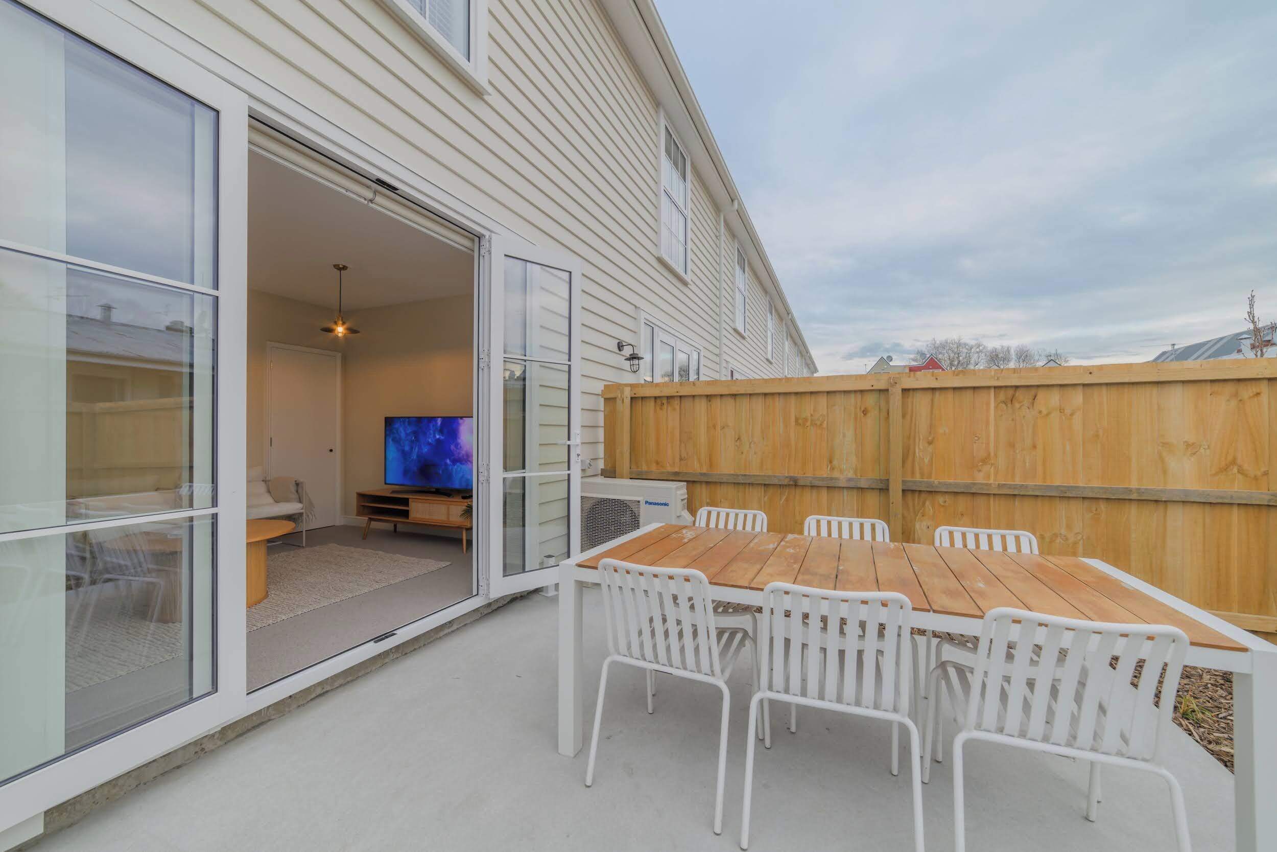 A patio with a wooden table and white chairs, a television in the background, and a wooden fence.