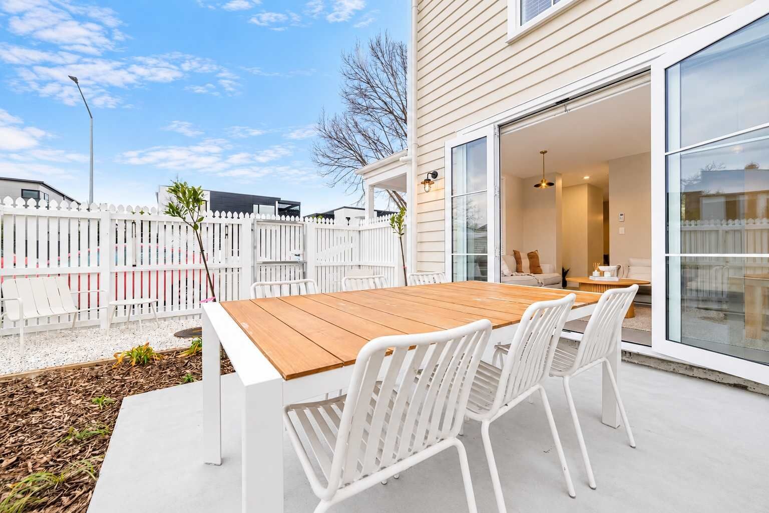A patio with a wooden table and white chairs, a white fence, and a house with large windows.