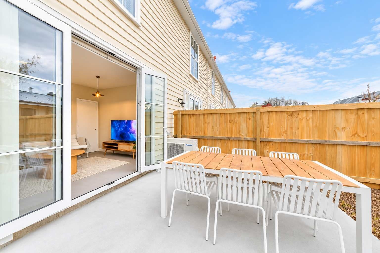 An outdoor patio with a wooden table and white chairs, a sliding glass door leading to an interior space with a TV, and a wooden fence.