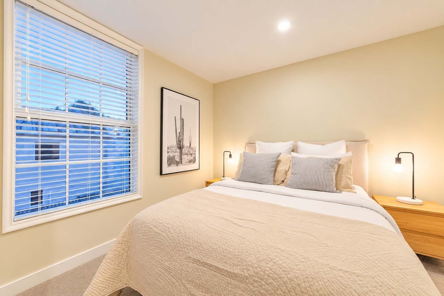 A bedroom with a bed, bedside lamps, a framed picture on the wall, and a window with blinds.