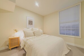 Un dormitorio con cama, una mesita de noche con lámpara, una ventana con persianas y una obra de arte colgada en la pared.