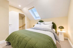 A bedroom with a skylight, a bed with green bedding, a bedside lamp, and a door leading to another room.