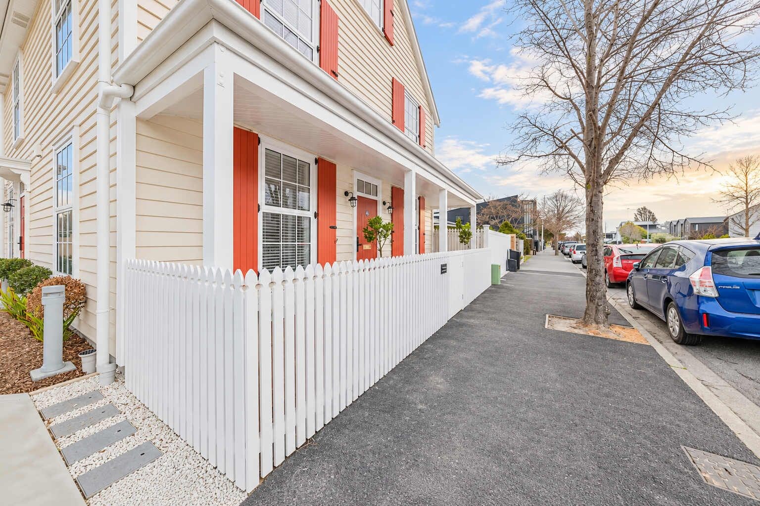A residential street with a white picket fence, a row of houses with red shutters, and parked cars.