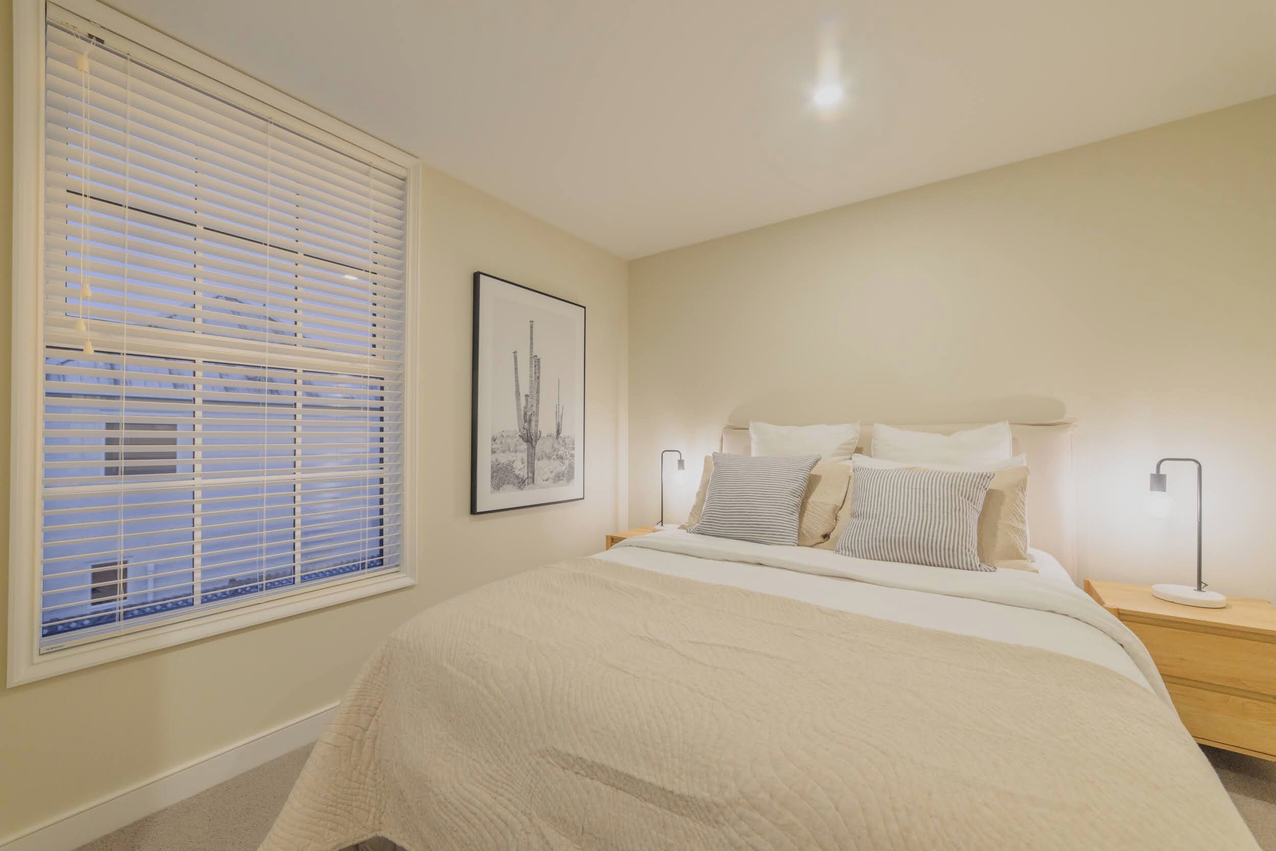 A bedroom with a bed, bedside tables, a window with blinds, and a framed picture on the wall.