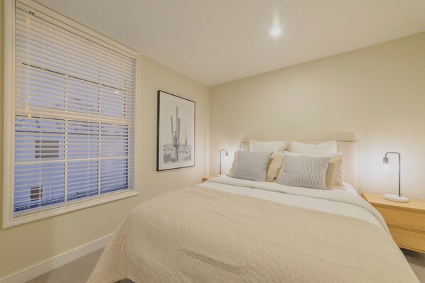 Un dormitorio con cama, mesitas de noche, una ventana con persianas y un cuadro en la pared.