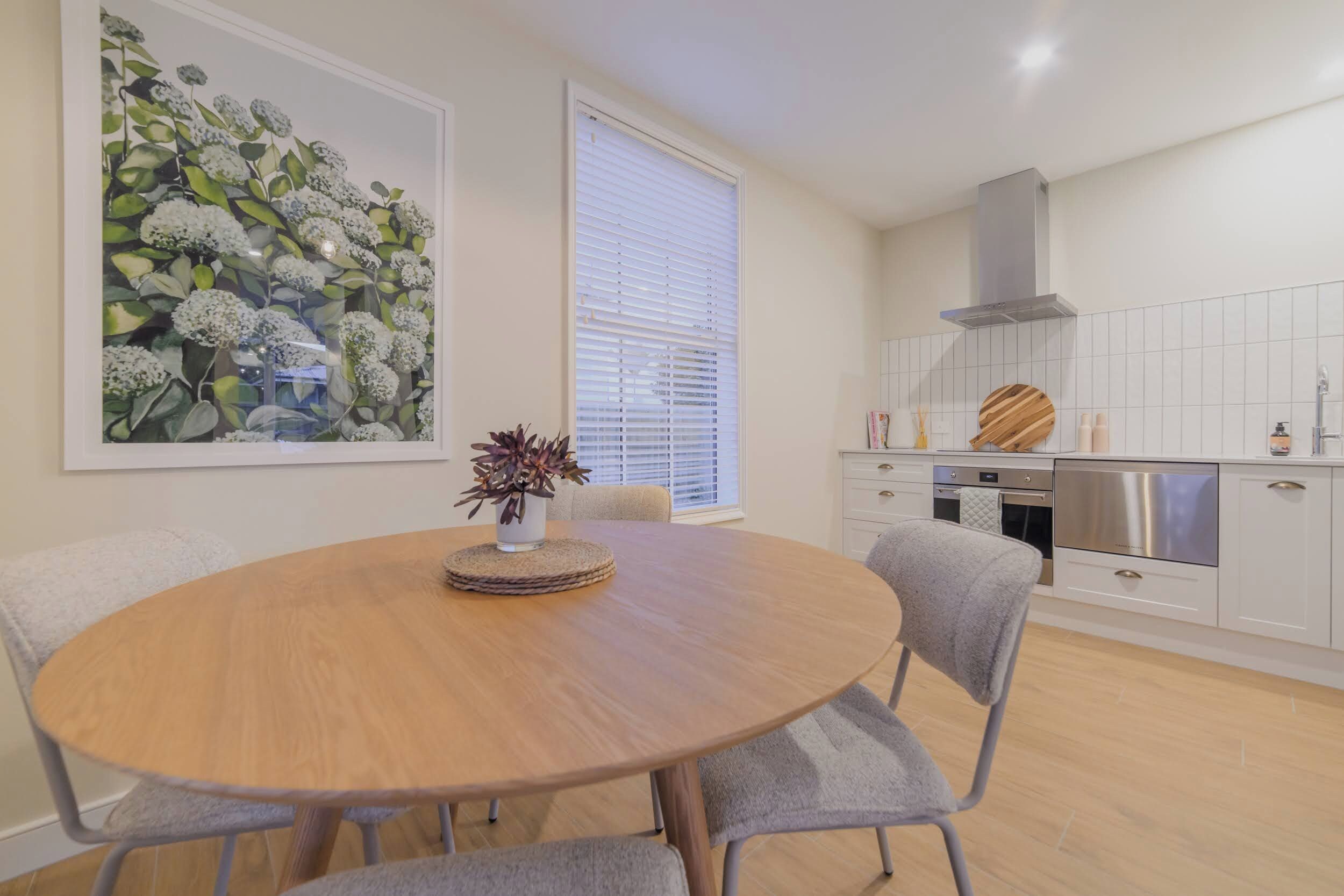 Un coin repas avec une table ronde en bois, des chaises à revêtement gris, une plante en pot et une grande œuvre d’art florale encadrée accrochée au mur.