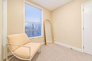 A room with a beige carpet, a white chair, a large window with blinds, a full-length mirror, and a white door.