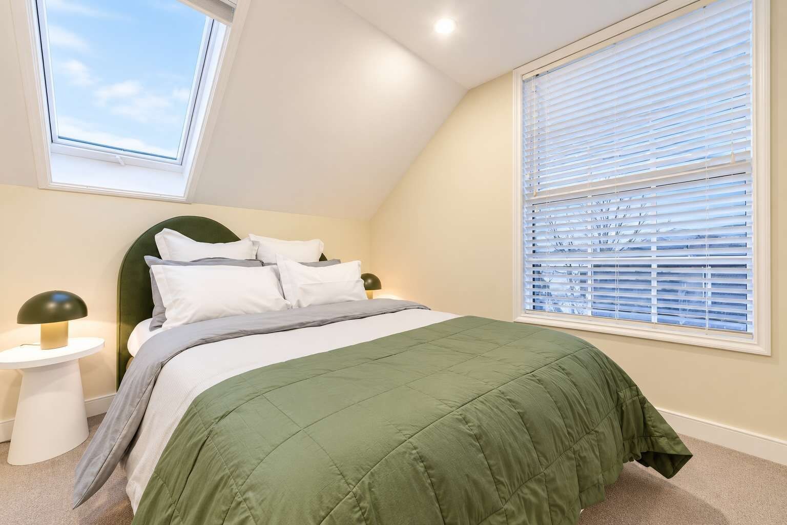 A bedroom with a bed, two bedside tables, a window with blinds, and a skylight.
