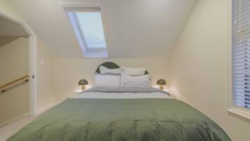 A bedroom with a skylight, a bed with a green bedspread, two bedside tables with lamps, and a window with blinds.