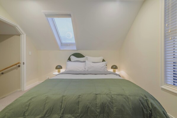 A bedroom with a skylight, a bed with a green bedspread, two bedside tables with lamps, and a window with blinds.